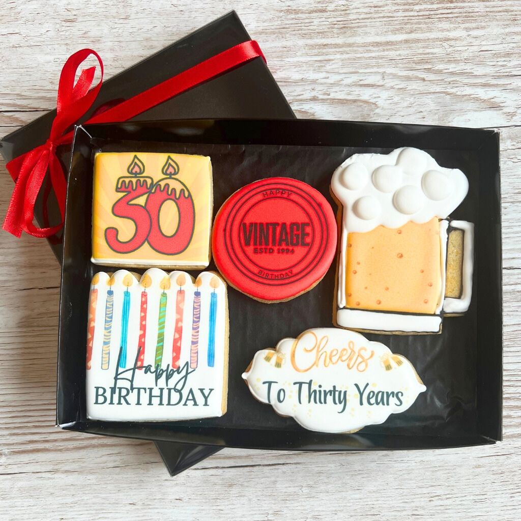 A boxed birthday cookie set displayed on a light wood surface, with a black gift box and red ribbon. The cookies are shaped like a yellow 50th birthday cake topper, a red vintage seal, a mug of beer with white foam, colorful birthday candles, and a white cloud-shaped cookie reading 'Cheers To Thirty Years'.